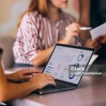 A group of professionals engaged in a business meeting, focused on data analysis displayed on a laptop screen. Charts and graphs indicate an analytical approach to teamwork and collaboration.
